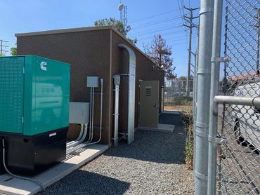A generator and electrical wiring outside of a pre-built building protected by a chained link fence.