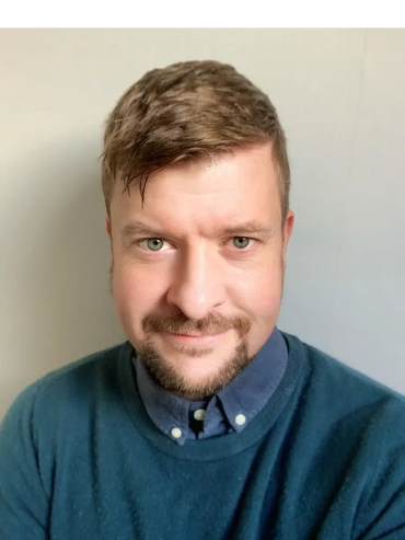 Smiling man with light brown hair and green eyes wearing a blue sweater and collared shirt.