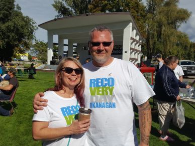 Smiling couple wearing matching 'Recovery Day Okanagan' shirts at an outdoor event.