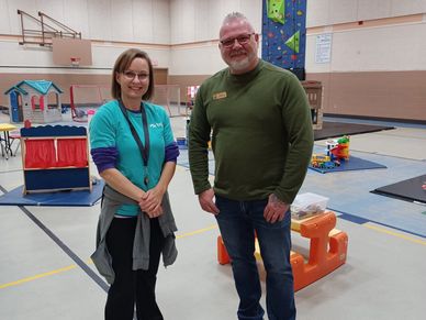 Two adults smiling inside a gymnasium with children's play equipment.