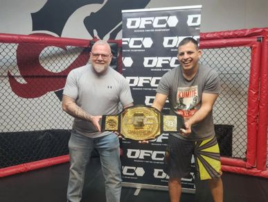Two men holding a championship belt inside a cage with DFC branding.