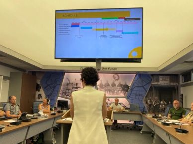 Person in white speaking at a conference room with a schedule on screen.