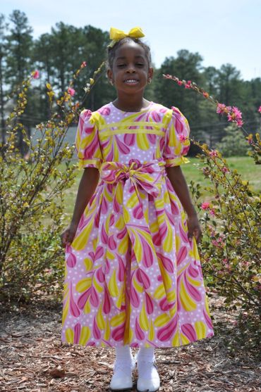 A smiling girl in a vibrant pink and yellow dress with a matching bow.