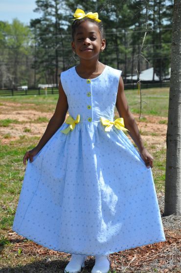 Young girl wearing a blue dress with yellow bows and a matching hair bow outdoors.