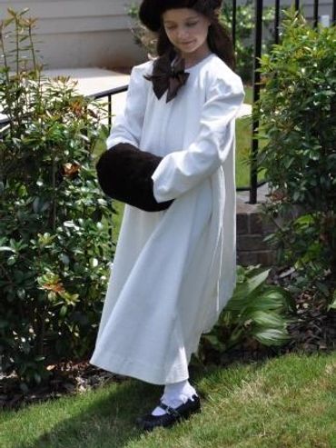 A young girl dressed in a white vintage outfit with fur accessories stands in a garden.