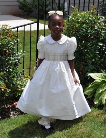 Young girl in a white dress standing on grass near bushes.