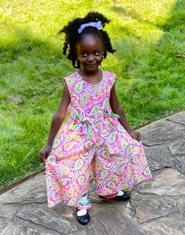 Smiling girl in a colorful paisley jumpsuit standing outdoors on a stone pathway.