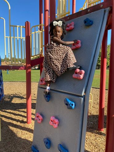 A girl in a giraffe-print dress climbing a playground rock wall on a sunny day.
