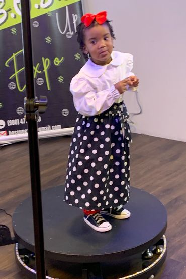A young girl in a white blouse and polka-dot skirt stands on a platform with a red bow in her hair.
