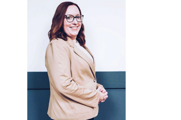 Smiling woman in beige blazer and glasses posing against a two-tone wall.
