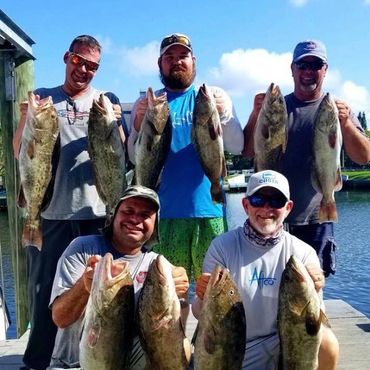Five men proudly display their large catches by the water on a sunny day.