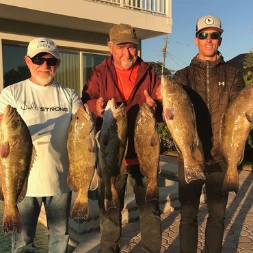 Three men proudly display their large catches of fish outdoors at sunset.