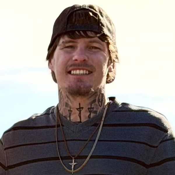 Young man with tattoos and necklaces smiling, wearing a backward cap.