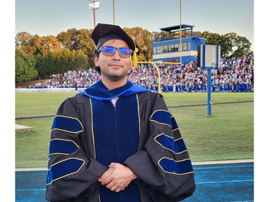Graduate in academic regalia standing on a sports field with a crowd in the background.