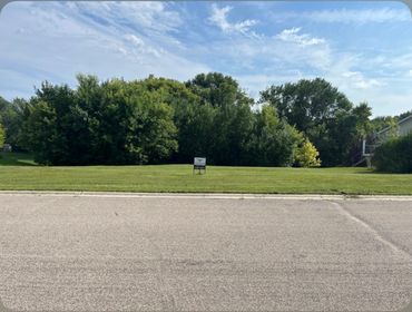 A grassy plot of land with a "For Sale" sign and trees in the background.