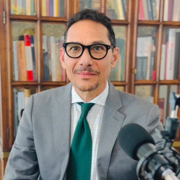 Man in suit and glasses speaking into microphone with bookshelf background.