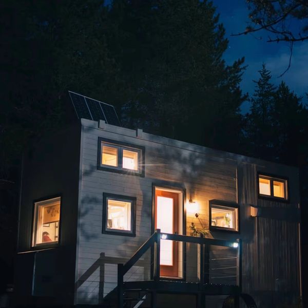 Cozy tiny house lit warmly at night amidst dark forest surroundings.