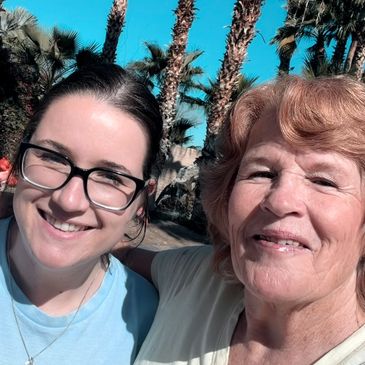 Two women smiling together outdoors under palm trees.