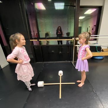 Two young girls play tic-tac-toe with toilet paper rolls on a dance studio floor.