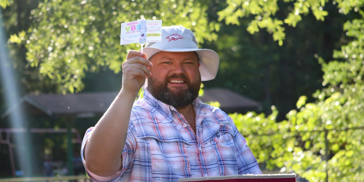 Man smiling while holding a small sign outdoors.