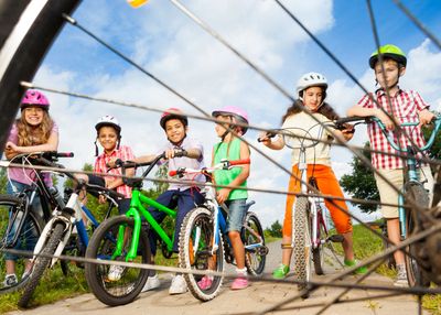 Looking through the spokes of a bike wheel, there is a lineup of children wearing helmets while on b