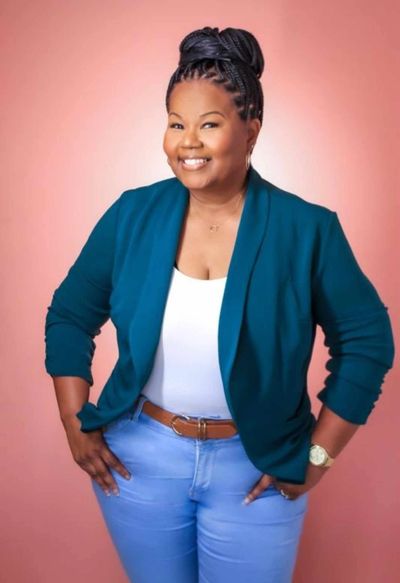 Smiling woman in a teal blazer and jeans against a pink background.