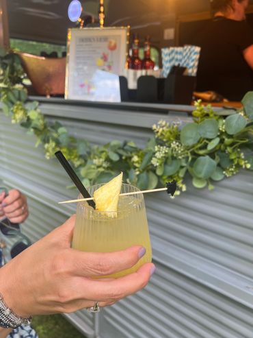 Hand holding a tropical cocktail with pineapple garnish at an outdoor bar.