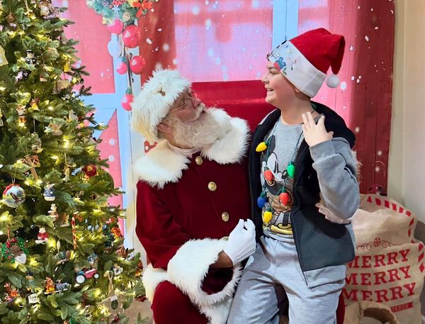 Santa Claus talks with a smiling child beside a decorated Christmas tree.