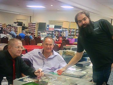 Three men at a table displaying coin collections in a busy room.