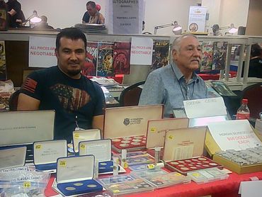 Two men sit behind a table selling collectible coins at a convention.