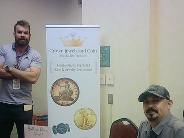 Two men at a Crown Jewels and Coin booth with flash sale sign and jewelry display.
