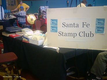 Santa Fe Stamp Club table at a stamp collecting event with members and materials.