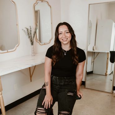 Young woman with long hair smiles, seated on a stool in a stylish room with mirrors and a marble countertop.