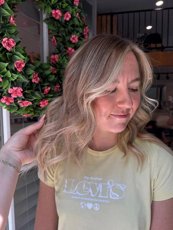 Woman with styled blonde hair and a yellow Levi's t-shirt stands near a floral wreath.