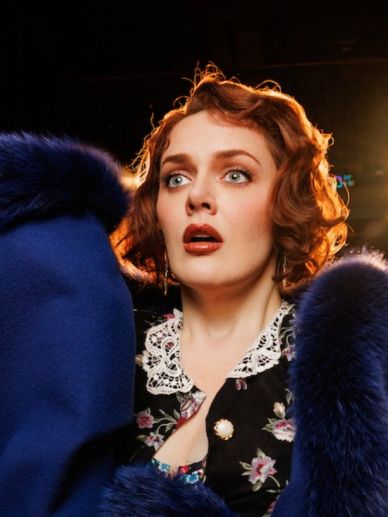 A woman with red hair and a blue fur coat looks surprised on a dimly lit stage.