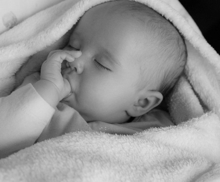 An infant, swaddled in a soft white blanket, sleeps while sucking her thumb.