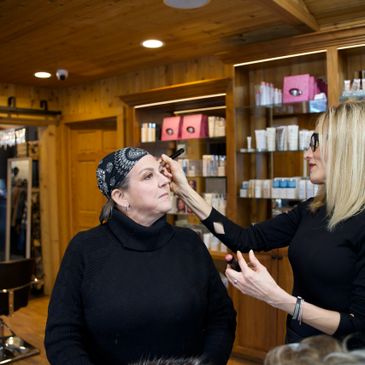 A makeup artist applies makeup on a seated woman in a wooden interior.
