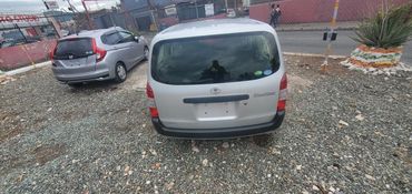 Rear view of a silver Toyota Probox parked on a gravel lot beside a silver Honda car.