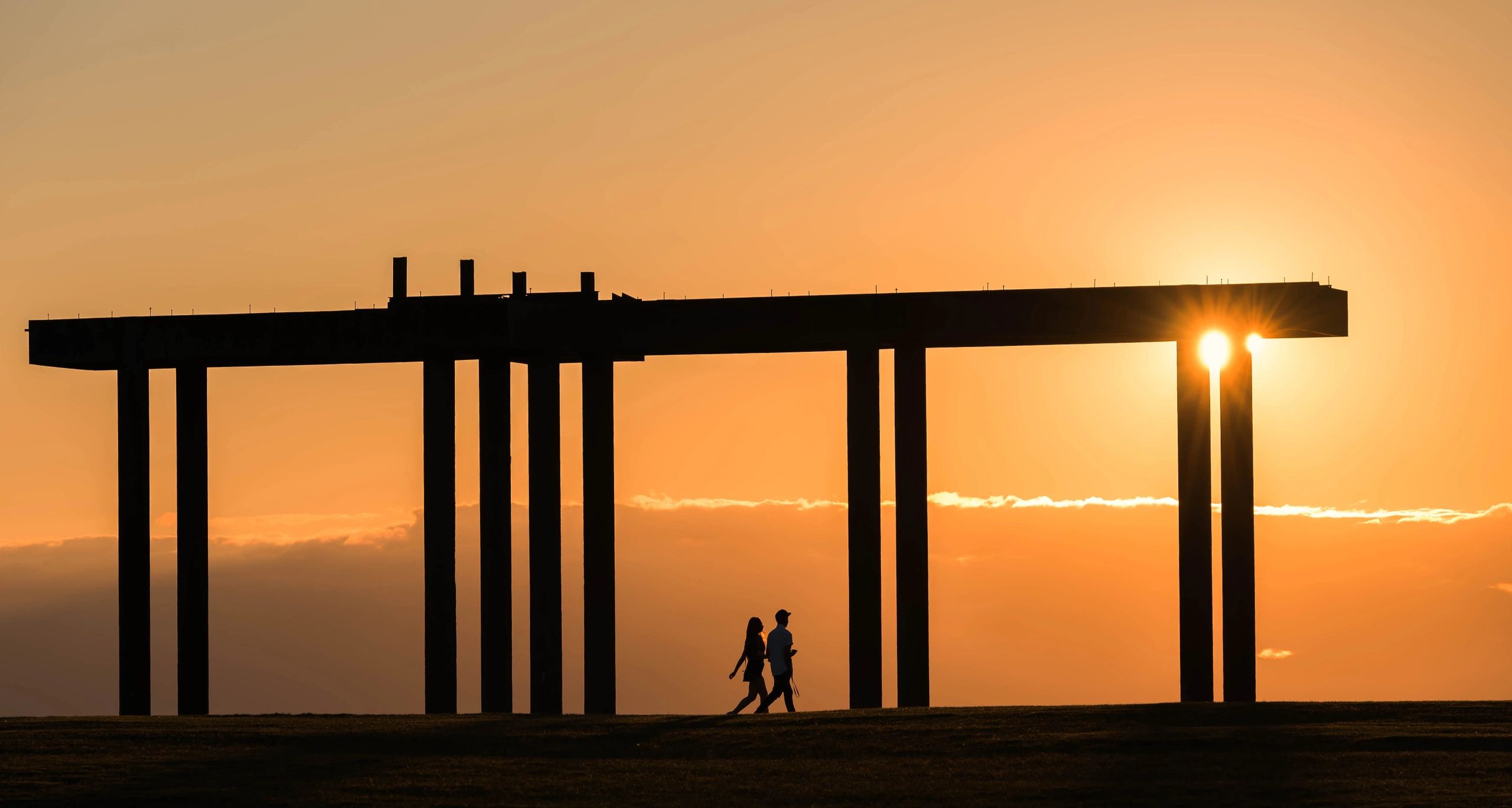Silhouetted couple walking near a bridge structure at sunset.