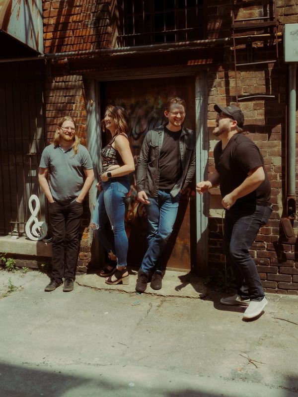Four friends casually hanging out in a sunlit urban alleyway with brick walls.