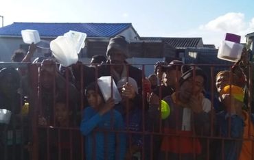 People stand behind a metal gate holding containers, possibly waiting for food or aid.