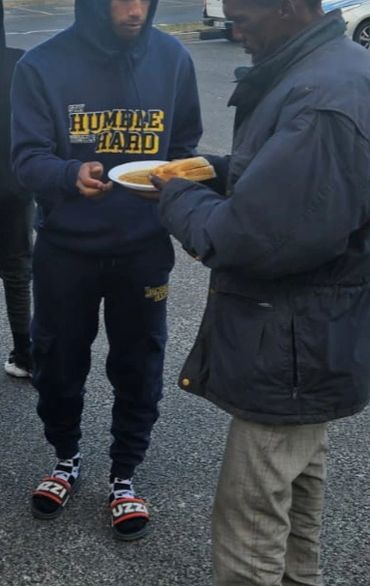 A man in a hoodie gives food to another man outside in a parking lot.