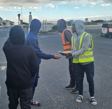 Four hooded individuals, two wearing reflective vests, interact on a street corner.