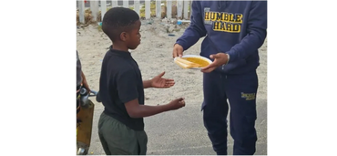 A boy receiving food from a person wearing a 'Humble Hard' outfit outdoors.