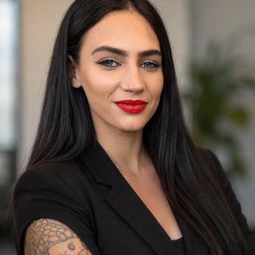 Confident woman with long dark hair and a tattoo, wearing a black blazer and red lipstick.