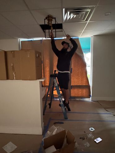 A man on a ladder installs a light fixture in a dimly lit room under renovation.