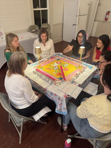 Six women play a traditional tile-based game around a decorated table.
