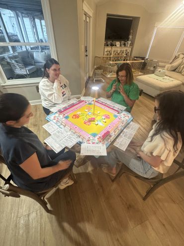 Four women playing a colorful board game around a table in a cozy living room.