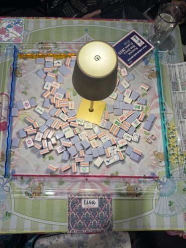 A mahjong game setup with tiles scattered inside a glass tray and a small lamp in the center.