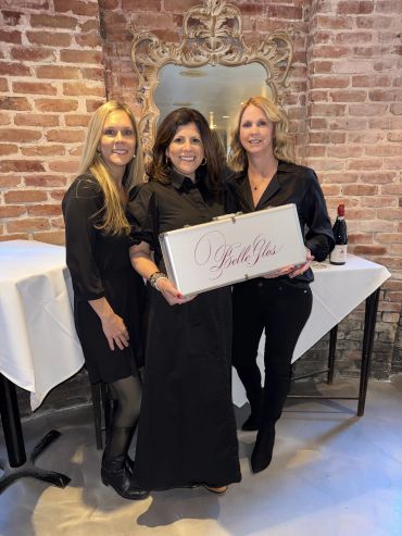 Three women dressed in black holding a Belle Glos case in a brick-walled room.